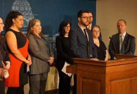 jay, a white man in a suit and glasses, speaking at a press conference at the capitol with a group of people