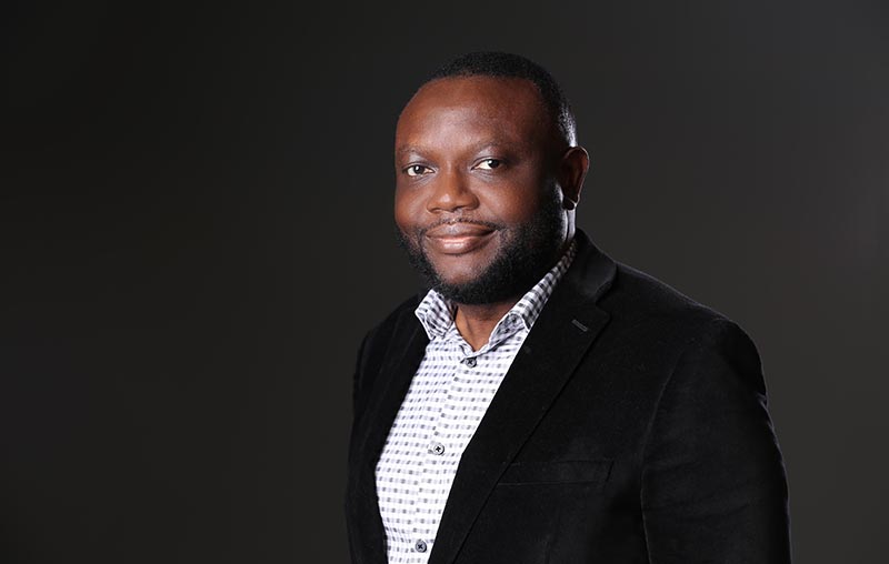 a black man smiles in front of a dark background in a suit coat