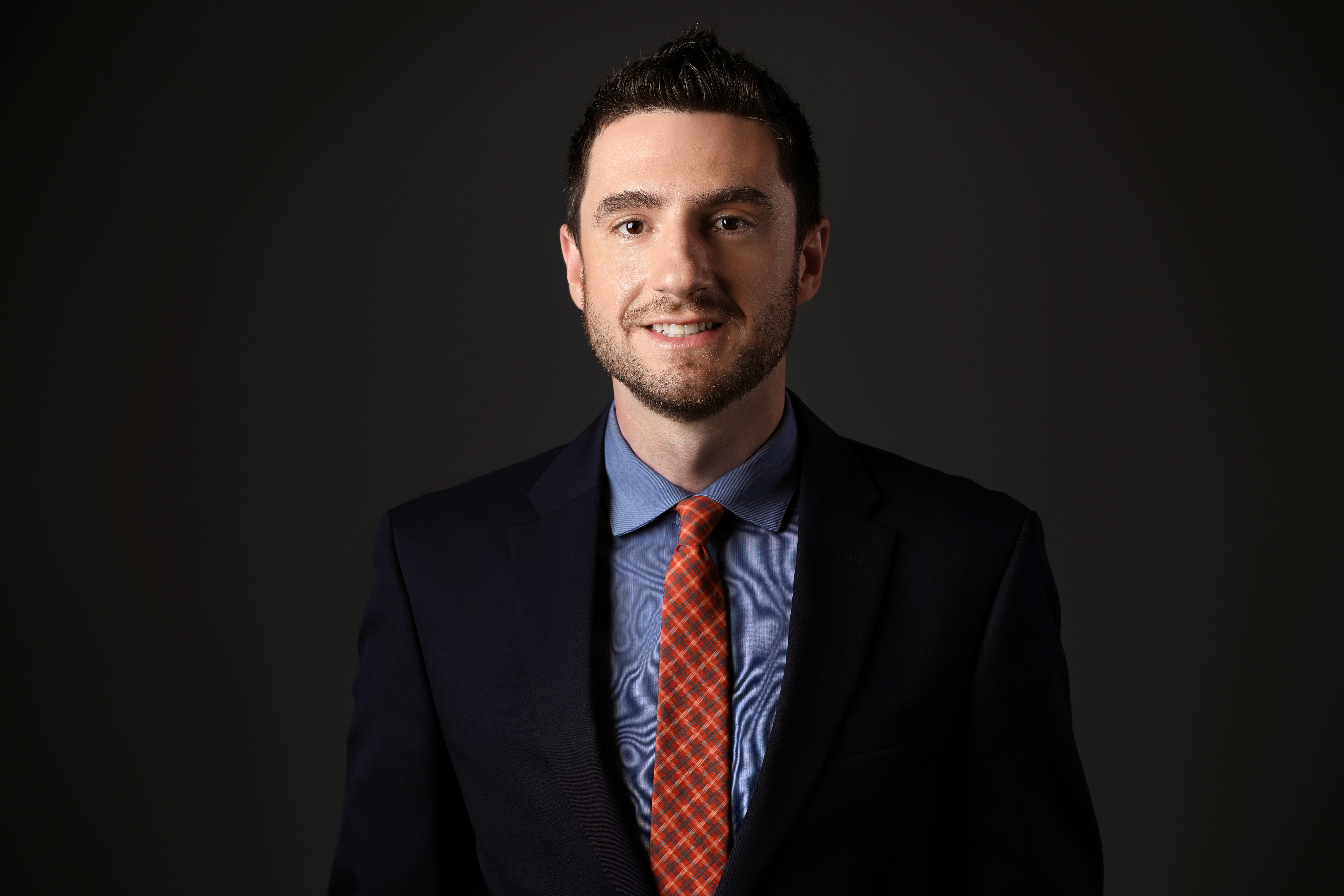 a white man in a suit coat smiles in front of a dark background