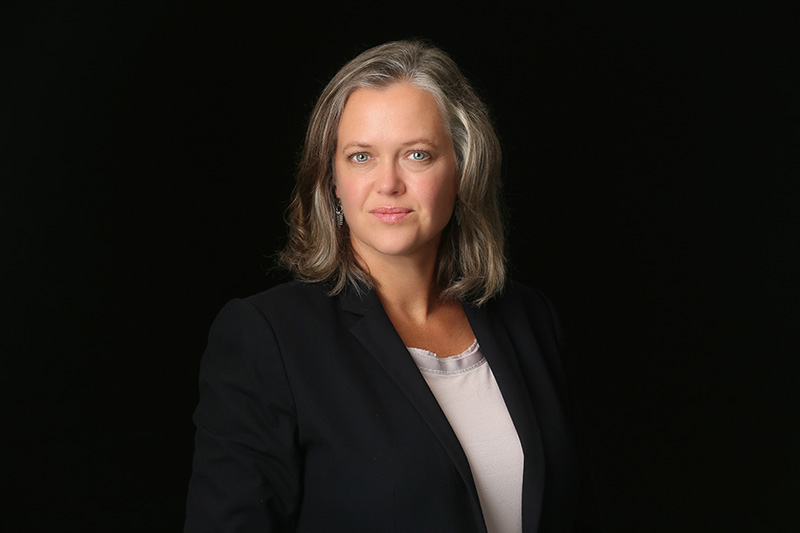 A white woman with silver hair in front of a dark background with a white shirt and black jacket 