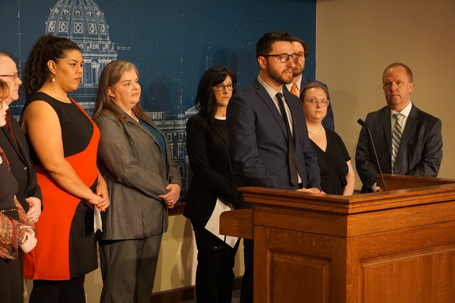 jay eidsness, a white man in a suit and glasses, stands speaking into a microphone with a group of people