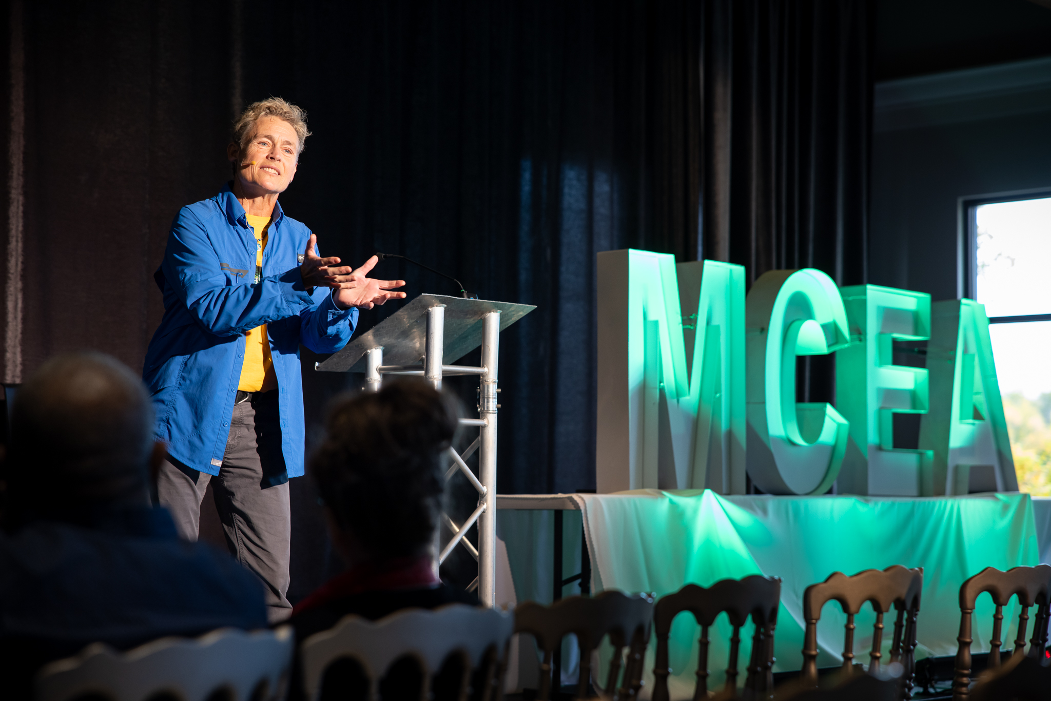 a white woman with short hair in a blue shirt holds out her hands in front of an m c e a logo