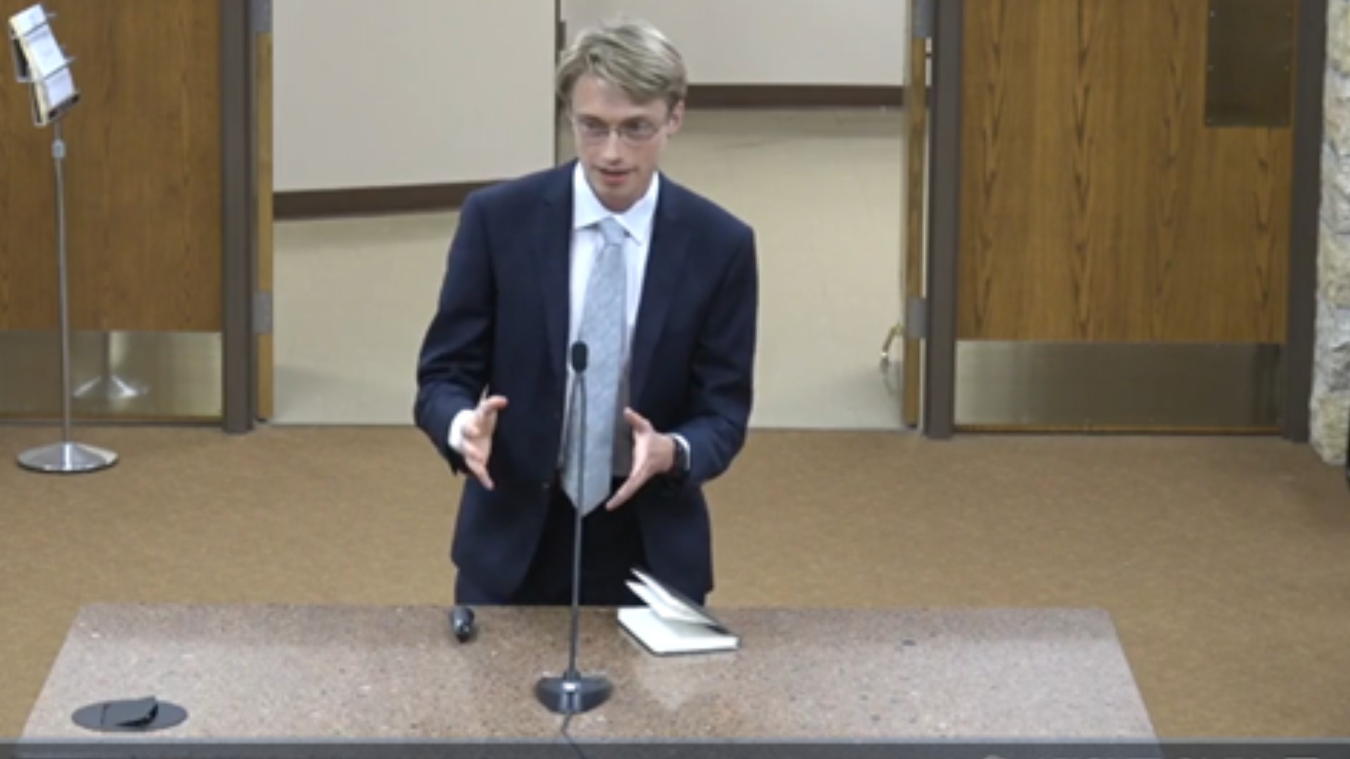 luke, a young white man in a suit testifies behind a brown table