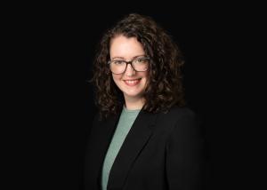 a white woman with curly hair smiles, wearing a blazer against a dark background