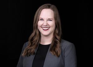 a white woman with long brown hair smiling widely in a gray jacket on a dark background
