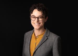 a photo of a white woman with short curly hair in a yellow shirt and gray suitcoat, smiling, on a dark background