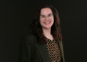 A photo of a white person with long, brown hair, glasses, a brown diamond printed shirt, and a green blazer. They are smiling widely. 