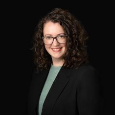 a white woman with curly hair smiles, wearing a blazer against a dark background