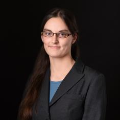 a white woman with long hair in a dark suitcoat standing in front of a dark background