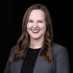 a white woman with long brown hair smiling widely in a gray jacket on a dark background