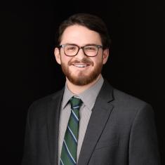 a white man with a beard, smiling, standing in a gray suit and tie