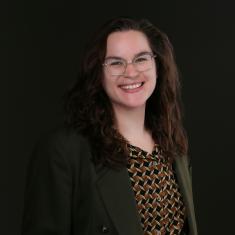 A photo of a white person with long, brown hair, glasses, a brown diamond printed shirt, and a green blazer. They are smiling widely. 