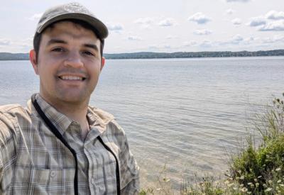 a photo of a white man in a hat smiling in front of a lake
