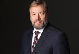 photo of Aaron Klemz, a white man with a beard wearing a striped tie and suit coat in front of a dark background