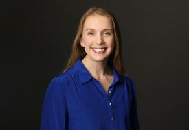 a smiling white woman with long straight hair in a blue button down shirt in front of a dark background