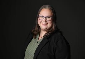 a photo of Andrea Lovoll, a white woman in glasses, with a bright smile in a green shirt and suit coat, against a dark background