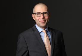 a photo of Evan Mulholland, a white man smiling slightly against a dark background in a suit and tie