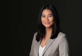 Photo of a Mainhia Thao, an East Asian woman with long black hair, in a gray suitcoat and white v neck on a dark background