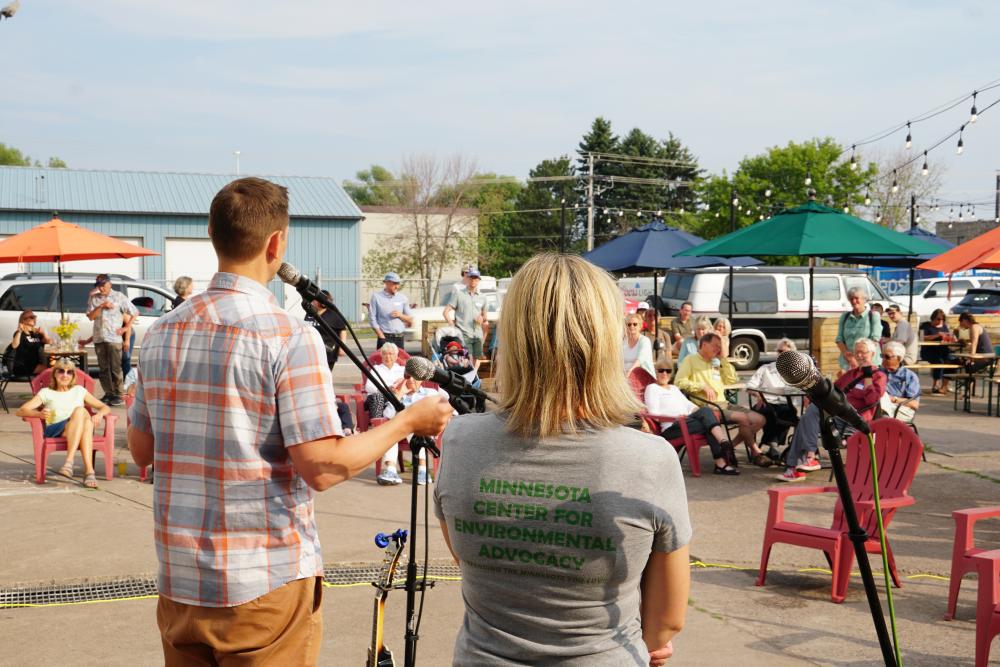 Photo of two M C E A staff with their backs to the camera speaking to the crowd of supporter at the M C E A event at Wild State Cider