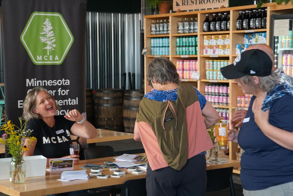 photo of M C E A staff member seated at a table waving at a supporter approaching